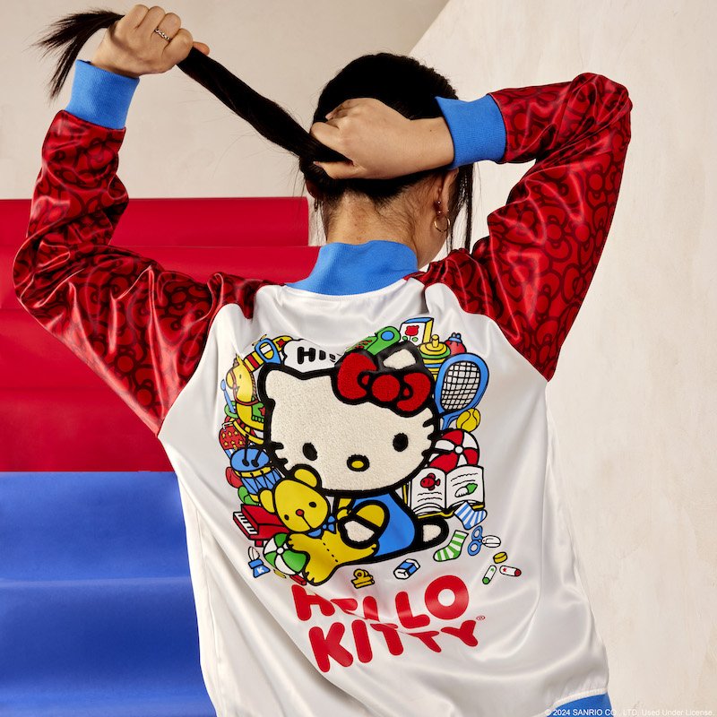 Woman facing away from camera standing on blue and red stairs and wearing the Sanrio Hello Kitty 50th Anniversary Unisex Souvenir Jacket. She's pulling her hair up into a ponytail to reveal the Hello Kitty design on the back of the jacket 